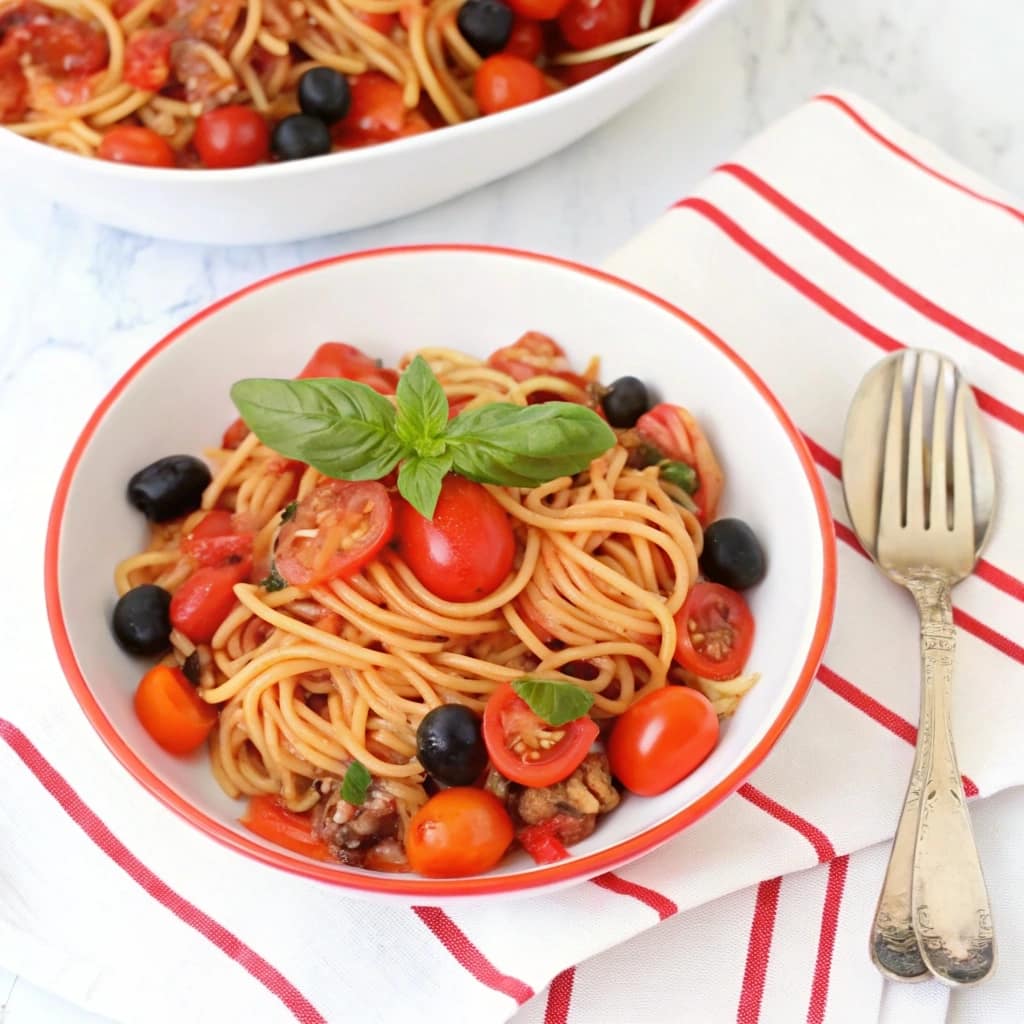 A bowl of vegan tomato basil lentil pasta with spaghetti, red peppers, and olives, garnished with fresh basil.