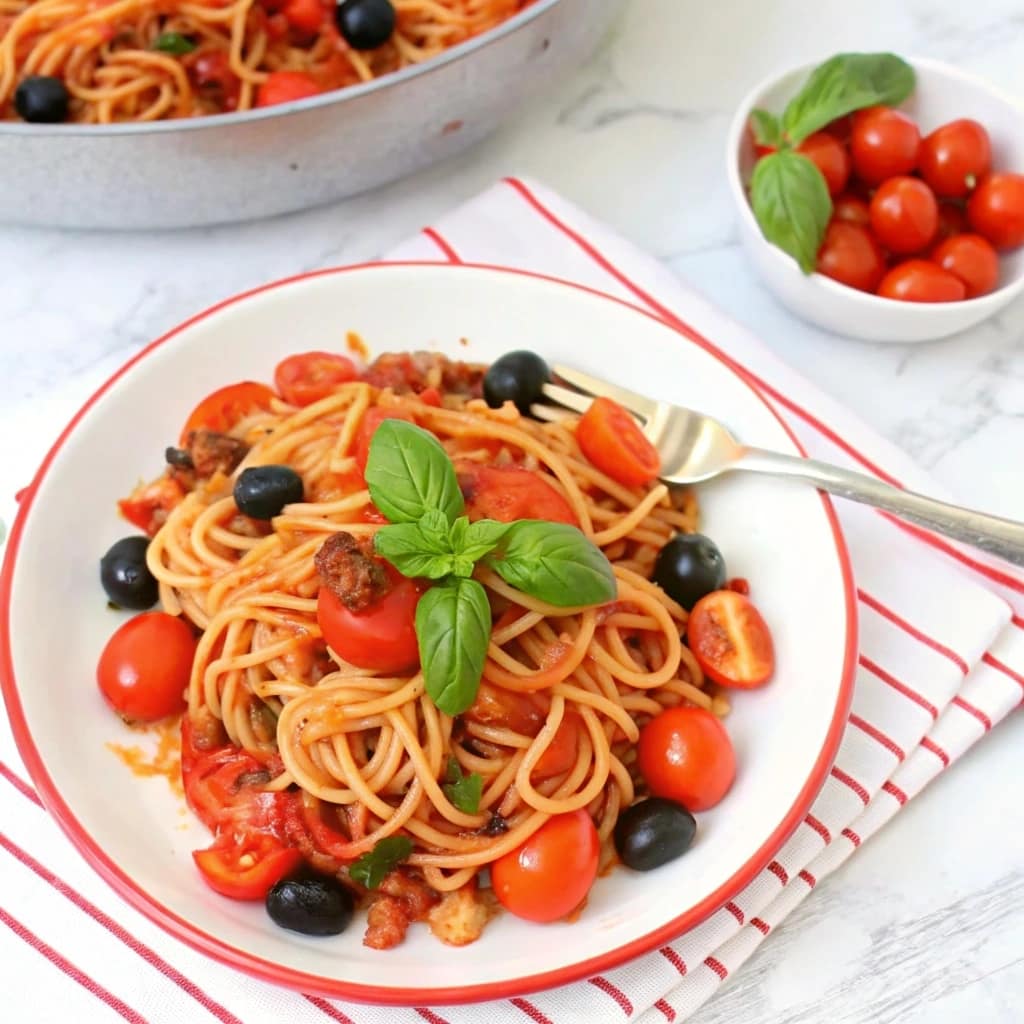 A bowl of vegan tomato basil lentil pasta with spaghetti, red peppers, and olives, garnished with fresh basil.