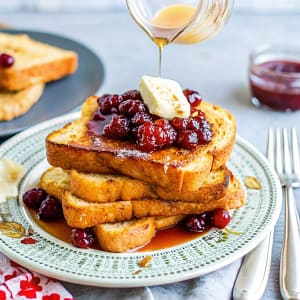 Golden brown eggnog French toast on a white plate with a pat of orange butter melting on top, garnished with an orange slice.