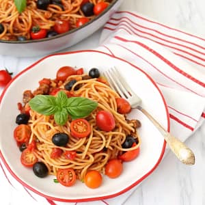 A bowl of vegan tomato basil lentil pasta with spaghetti, red peppers, and olives, garnished with fresh basil.