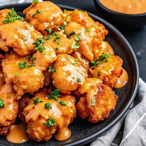 A close-up of crispy golden Bang Bang chicken tenders coated in a creamy white and red sauce, garnished with parsley, on a dark plate.